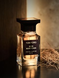 Ein Parfümflakon „Bois Marocain“ von Tom Ford steht auf einer dunklen Fläche neben getrocknetem Gras, während von hinten warmes Licht darauf fällt.
