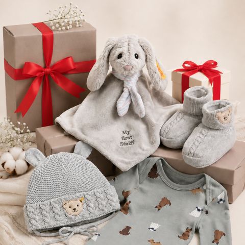 Baby-Geschenkset mit Plüschhäschen, grauer Strickmütze, grauen Babyschuhen, einem Strampler mit Bärenmotiv und verpackten Geschenken mit roten Schleifen auf einem beigen Hintergrund.