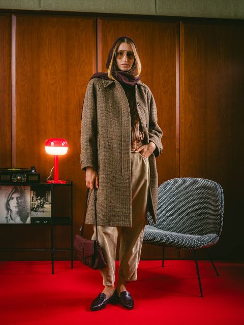 Eine Person in einem braunen Mantel und einem Schal steht in einem Raum auf einem roten Teppich, im Hintergrund sind Vintage-Dekorationen, ein Stuhl und eine rote Lampe zu sehen.
