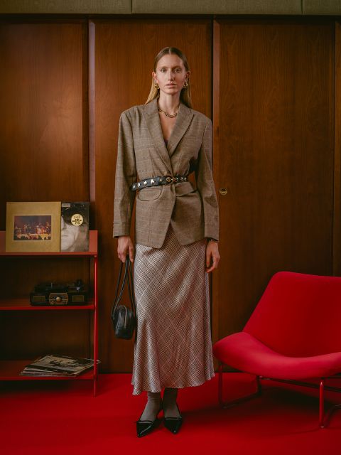 Eine Frau in Blazer und kariertem Rock steht in einem Retro-Zimmer mit holzvertäfelten Wänden, rotem Teppich, rotem Stuhl und Gegenständen auf einem roten Regal.