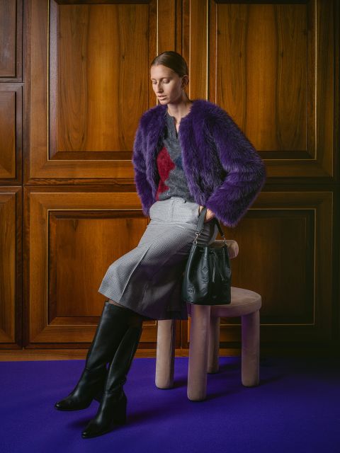 Eine Frau in einer violetten Pelzjacke, einem grauen Rock und schwarzen Stiefeln sitzt mit einer schwarzen Handtasche auf einem Hocker vor Holzvertäfelungen auf einem violetten Teppich.