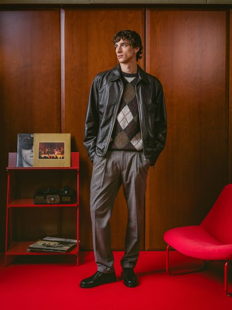 Ein Mann in einer Lederjacke und einem karierten Pullover steht auf einem roten Teppich neben einem roten Sessel, einem Retro-Radio und Alben vor holzvertäfelten Wänden.
