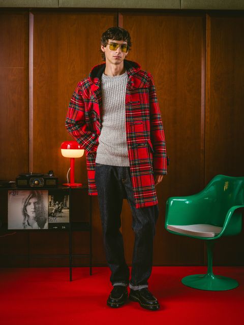 Eine Person in einem roten Karomantel, einem grauen Pullover, dunklen Jeans und einer Sonnenbrille steht in einem Raum im Retro-Stil mit Holzvertäfelung, einem grünen Stuhl und einer roten Lampe.