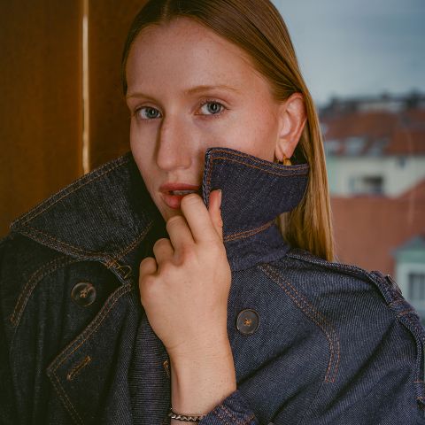 Eine Frau mit heller Haut und glattem Haar, die eine dunkle Jeansjacke trägt, deren Kragen sie hochgestellt hat, und vor einem Fenster steht, im Hintergrund sind Gebäude zu sehen.