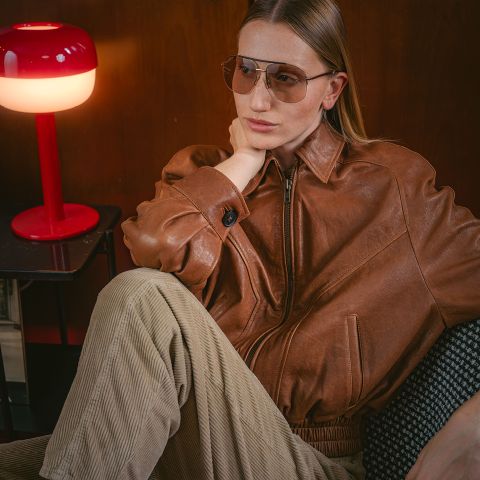 Eine Person in einer braunen Lederjacke und einer Pilotenbrille sitzt neben einer roten Lampe und wirkt nachdenklich, während ihre Hand auf dem Nacken ruht.