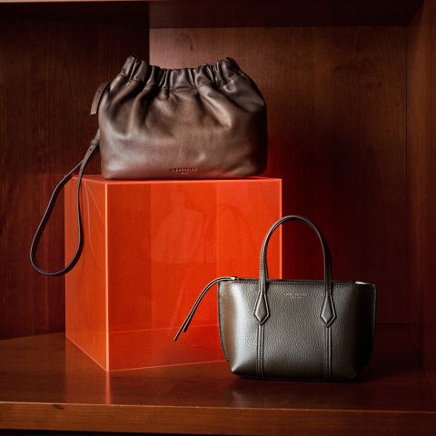 Zwei elegante Lederhandtaschen, eine dunkelbraune und eine olivfarbene, präsentiert auf einem Holzregal zusammen mit einem orangefarbenen, durchscheinenden Würfel.