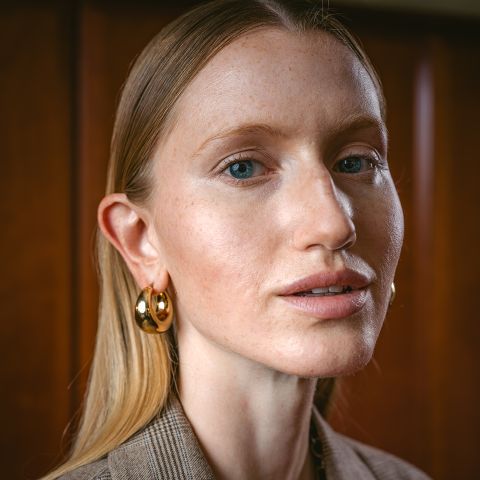 Eine Frau mit glattem blondem Haar, goldenen Creolen und dezentem Make-up blickt in die Kamera, während sie vor einem Holzhintergrund steht.