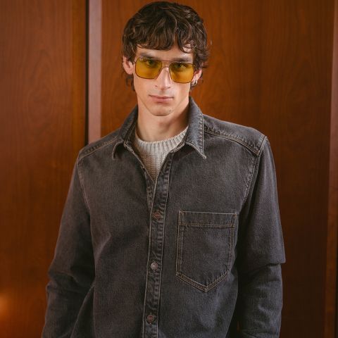 Ein junger Mann mit welligem braunem Haar, der eine gelb getönte Brille, ein graues Jeanshemd und einen weißen Pullover trägt und vor einem mit Holz vertäfelten Hintergrund steht.