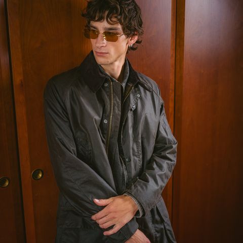 Man wearing a dark jacket and amber-tinted sunglasses stands with arms crossed, against wood-paneled doors, looking to the side.