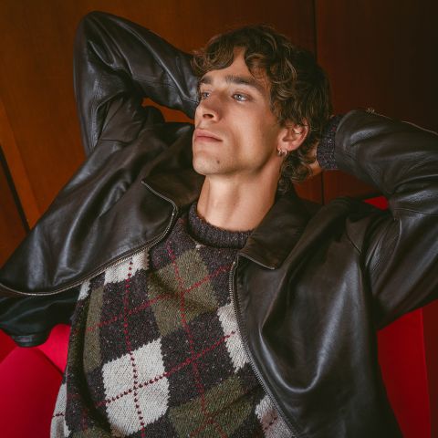 Eine Person in einer Lederjacke und einem Argyle-Pullover lehnt sich in einem roten Sessel zurück, die Hände hinter dem Kopf verschränkt, und blickt nachdenklich nach oben.