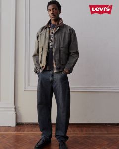 Ein Mann in weiten Jeans, einem T-Shirt mit Aufdruck und einer Jacke steht in einem Raum auf einem Holzboden. In der rechten oberen Ecke ist das Levi’s-Logo zu sehen.