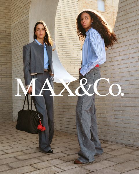 Zwei Frauen posieren in stilvollen Outfits von MAX&Co. vor einer Backsteinmauer; die eine trägt einen Anzug und eine Tasche, die andere ein gestreiftes Shirt und weite Jeans.