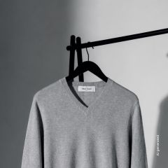Ein hellgrauer Pullover mit V-Ausschnitt hängt an einem schwarzen Kleiderbügel vor einem schlichten Hintergrund, teils im Schatten, teils im Licht.