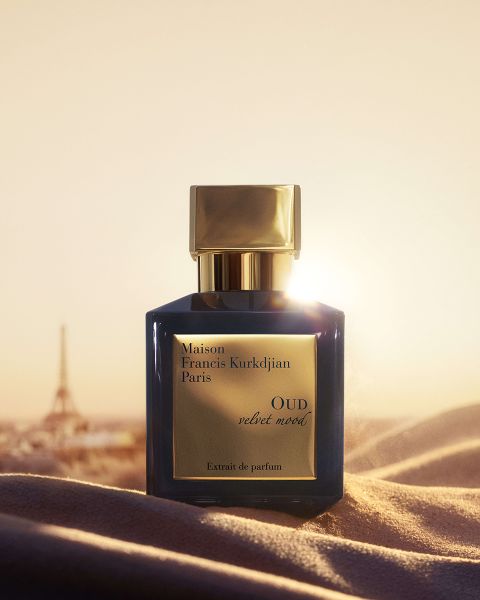 Ein blau-goldener Flakon des Parfüms „OUD Velvet Mood“ von Maison Francis Kurkdjian steht auf einem Stoff, im Hintergrund sind ein Sonnenuntergang und der Eiffelturm zu sehen.