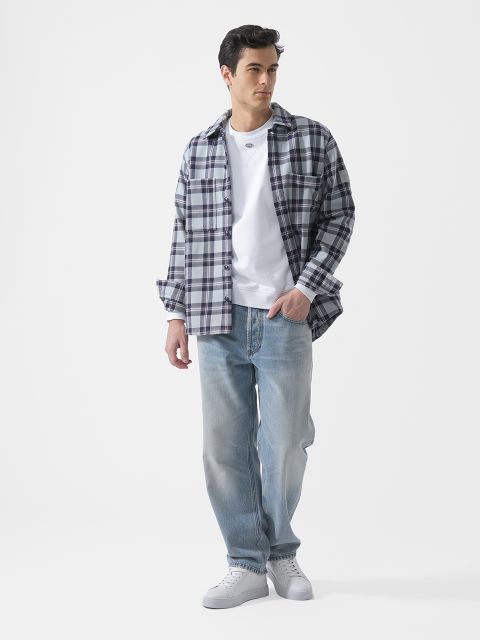 Ein Mann, der ein weißes T-Shirt, ein kariertes Hemd, hellblaue Jeans und weiße Turnschuhe trägt, steht vor einem schlichten weißen Hintergrund.