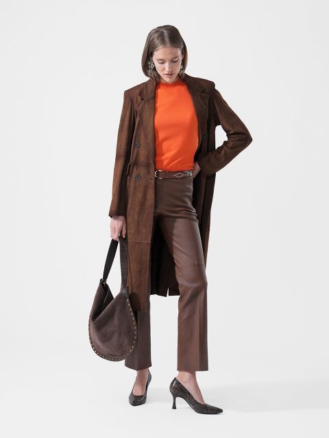 Eine Frau in einem braunen Mantel, einem orangefarbenen Oberteil, einer braunen Hose und Pumps hält eine braune Handtasche in der Hand und steht vor einem schlichten weißen Hintergrund.