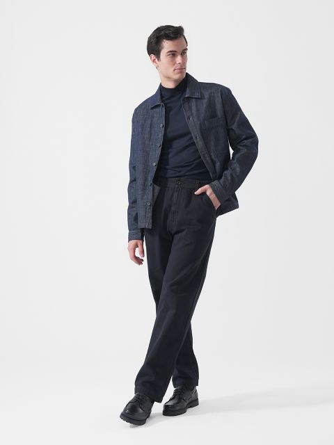 Ein Mann, der eine dunkle Jeansjacke, einen marineblauen Rollkragenpullover, eine schwarze Hose und schwarze Schuhe trägt, steht vor einem schlichten weißen Hintergrund und blickt nach links.