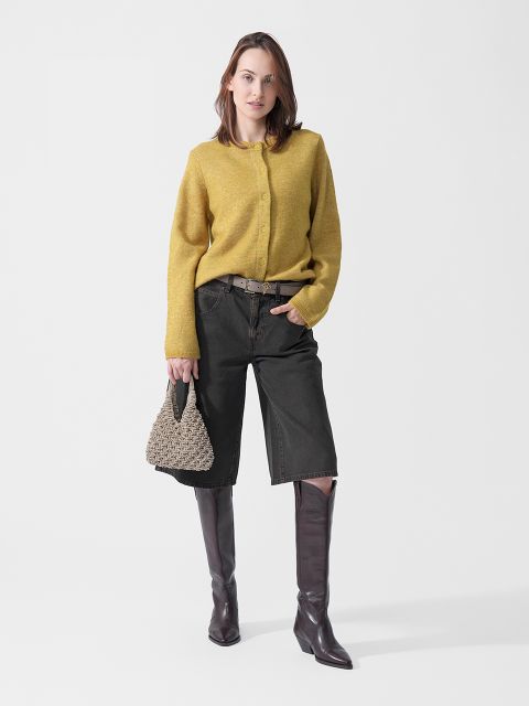 Eine Frau, die einen gelben Pullover, schwarze, knielange Shorts und braune Stiefel trägt und eine geflochtene Handtasche in der Hand hält, steht vor einem weißen Hintergrund.