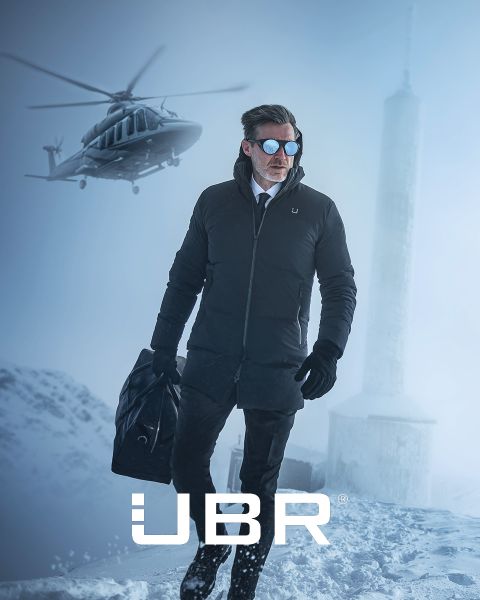 Ein Mann in einem schwarzen Mantel und mit Sonnenbrille steht im Schnee und hält eine Tasche in der Hand; im Hintergrund sind ein Hubschrauber und ein hoher Turm zu sehen. Das UBR-Logo ist zu erkennen.
