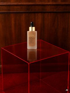 Auf einem durchsichtigen roten Acryltisch vor einer Holzwand und einem roten Teppich steht eine Flasche Estée Lauder-Serum.