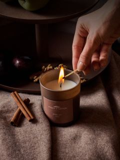 Eine Hand zündet eine braune „African Tea“-Kerze von Finch Hatton an, die neben Zimtstangen, Pflaumen und Walnüssen auf einem weichen braunen Tuch steht.