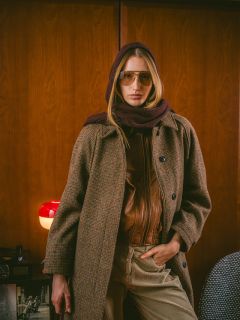 Eine Person mit Sonnenbrille, Schal und kariertem Mantel steht in einem Raum, eine Hand in der Tasche, im Hintergrund eine rote Lampe und Holzvertäfelung.