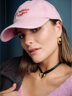 Eine Frau mit glattem braunem Haar trägt eine rosa Mütze, goldene Ohrringe, ein Herz-Halsband und ein rosa, flauschiges Oberteil und blickt vor einem blauen Hintergrund in die Kamera.