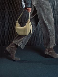 Eine Person in grauen Jeans und Wildlederstiefeln mit Absatz hält eine beige Handtasche in der Hand und geht vor einem strukturierten dunklen Hintergrund vorbei.