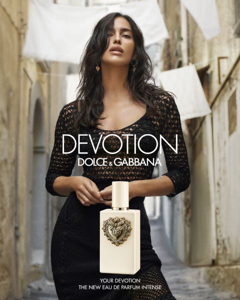 Eine Frau in schwarzer Kleidung steht in einer engen Gasse hinter einer Flasche „Dolce & Gabbana Devotion“-Parfüm. Im Hintergrund hängt weiße Wäsche.
