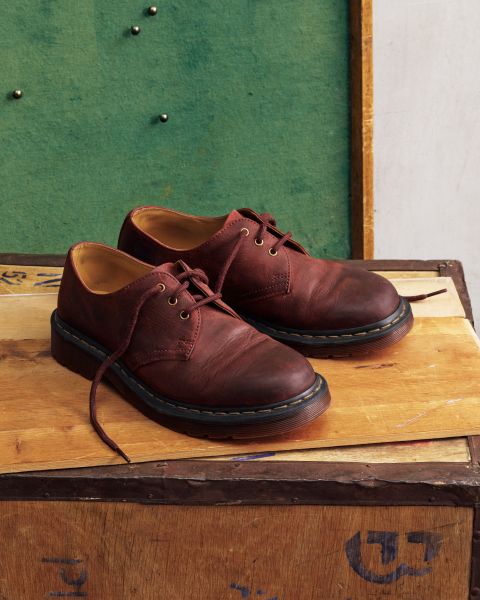 Ein Paar dunkelbraune Oxford-Schnürschuhe aus Used-Look-Leder mit gelben Nähten steht auf einer Holzfläche vor einer grünen Pinnwand.