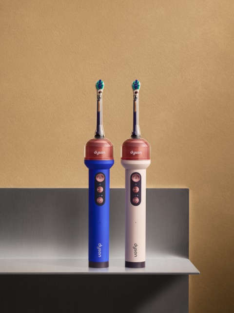 Two Dyson electric toothbrushes, blue and white, stand upright on a metal shelf against a beige wall.