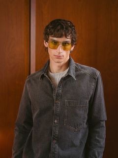 Ein Mann mit gewelltem Haar, der eine gelbe Sonnenbrille, ein graues Jeanshemd und einen hellen Pullover trägt, steht vor einer Holzwand und blickt in die Kamera.