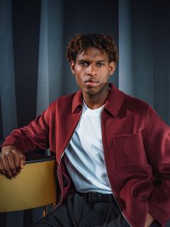 Ein Mann in einer kastanienbraunen Jacke und einem weißen Hemd sitzt neben einem goldenen Tisch und posiert vor dunklen Vorhängen.