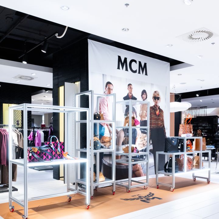 Storefront display in a mall with MCM banner and glass shelves filled with handbags.