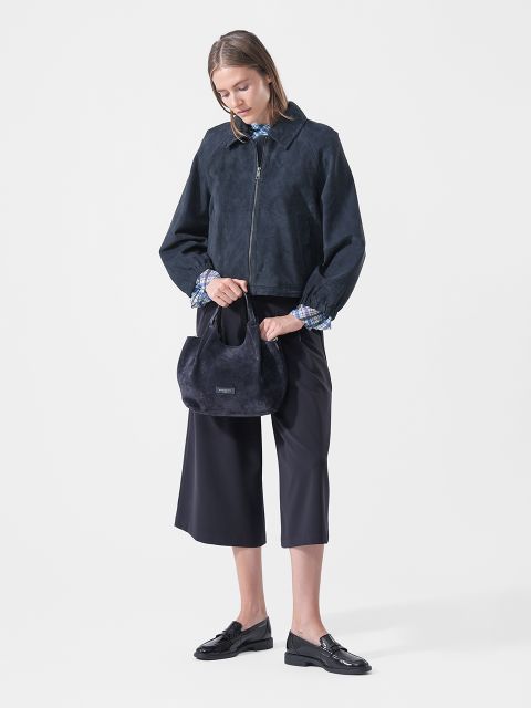 Eine Frau in einer marineblauen Jacke, einer Culotte und Loafern hält eine kleine marineblaue Handtasche in der Hand und steht vor einem schlichten weißen Hintergrund.