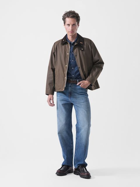 Vor einem schlichten weißen Hintergrund steht ein Mann mit dem Blick nach vorne, bekleidet mit einer braunen Jacke, einem Jeanshemd, blauen Jeans und schwarzen Schuhen.