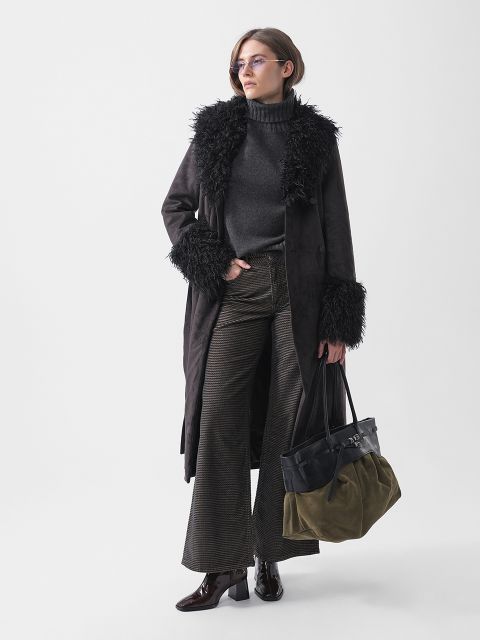 Eine Frau steht da, gekleidet in einen dunklen, mit Pelz besetzten Mantel, einen Rollkragenpullover, eine Schlaghose und Stiefel. Sie trägt eine zweifarbige Tasche und eine Brille und posiert selbstbewusst.