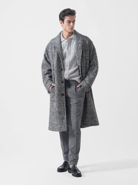 Ein Mann, der einen grauen, strukturierten Mantel, ein hellgraues Hemd mit Knöpfen, eine graue Hose und schwarze Schuhe trägt, steht mit den Händen in den Taschen vor einem weißen Hintergrund.
