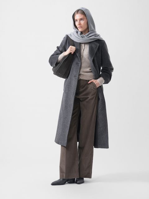 Eine Frau in einem grauen Kapuzenmantel, einem beigen Pullover, einer braunen Hose mit weitem Bein und schwarzen Schuhen, die eine schwarze Tasche in der Hand hält und vor einem weißen Hintergrund steht.