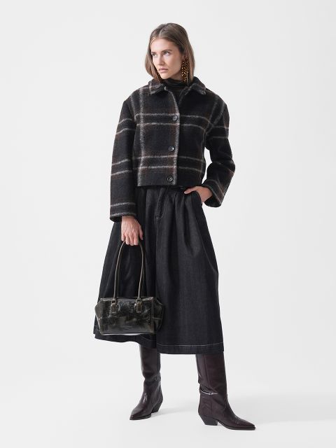 Eine Frau, die eine karierte Jacke, einen schwarzen Rock und hohe braune Stiefel trägt und eine schwarze Handtasche in der Hand hält, steht vor einem schlichten weißen Hintergrund.
