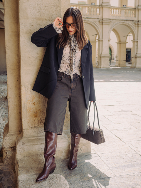 Eine Frau in einem Blazer, einer gemusterten Bluse und einer dunklen Jeans-Culotte, die kniehohe braune Stiefel trägt und eine Handtasche in der Hand hält, während sie in einem steinernen Innenhof steht.