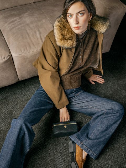 Eine Person in einer braunen Jacke mit Pelzkragen und Jeans sitzt auf einem Teppichboden, hält eine schwarze Clutch in der Hand und blickt mit ernstem Gesichtsausdruck nach oben.