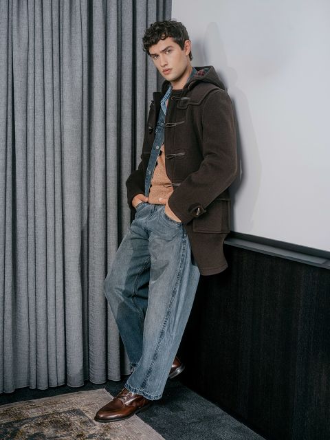 Ein junger Mann in einem braunen Mantel, einem orangefarbenen Pullover und blauen Jeans lehnt mit den Händen in den Taschen an einer Wand und steht neben grauen Vorhängen.