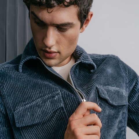 Ein Mann mit kurzen Locken, der eine blaue Cordjacke über einem weißen Hemd trägt, schaut nach unten, während er den Reißverschluss der Jacke schließt.