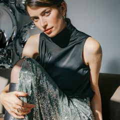 Eine Frau, die ein schwarzes, ärmelloses Oberteil mit Stehkragen und einen schimmernden grünen Paillettenrock trägt, sitzt auf einem Sofa.
