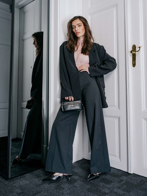 Eine Frau in einem schwarzen Blazer, einem rosa Spitzenoberteil und einer Hose mit weitem Bein hält eine kleine schwarze Handtasche in der Hand.