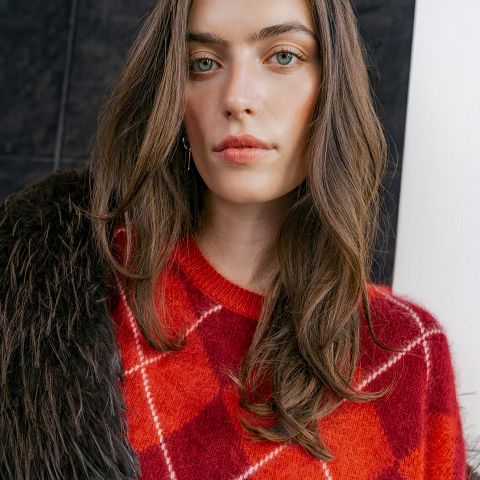 Eine Frau, die einen roten Argyle-Pullover trägt und eine dunkle Kunstfelljacke über die Schulter geworfen hat.
