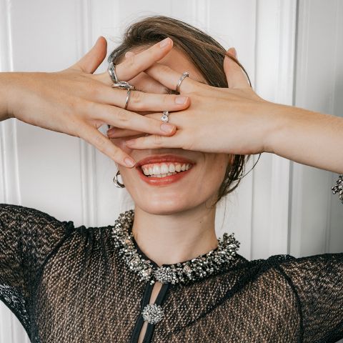 Eine Person, die lächelt und dabei die Hände vor die Augen hält, sodass mehrere silberne Ringe zu sehen sind.