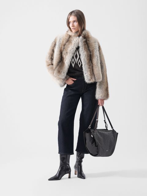 Ein Model trägt eine Pelzjacke über einem gemusterten Pullover, dunkle Jeans mit weitem Bein und schwarze Stiefel mit Absatz und hat eine große schwarze Umhängetasche dabei.