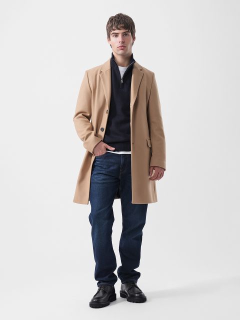 Ein Mann, der einen hellbraunen Mantel, einen marineblauen Pullover mit Reißverschluss am Kragen, ein weißes T-Shirt, dunkelblaue Jeans und schwarze Schuhe trägt.
